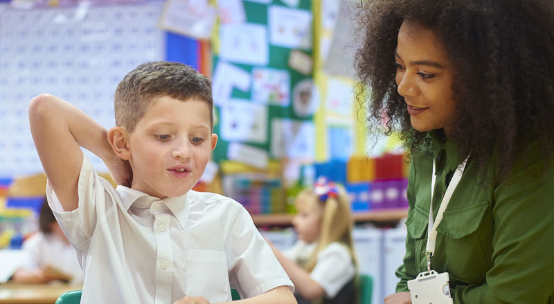 a teacher and a primary level student sitting at a desk in a brightly lit classroom. The both teacher and student are looking down at work on the desk and smiling.