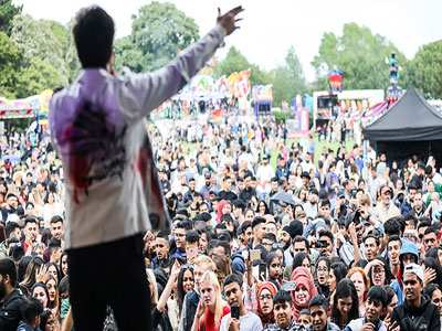 A photograph of a man singing from a stage. There is a large crowd in front of him and fairground rides are in the background.. A photograph of a man singing from a stage. There is a large crowd in front of him and fairground rides are in the background.. Link to Do Good, Feel Good - Middlesbrough Mela set up, Friday 15 August 2025.