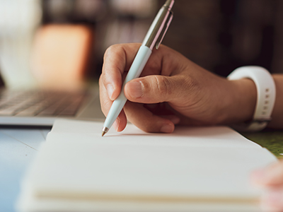 Person writing in a notebook (image shows hand, pen, notebook and laptop). Link to Develop your Essay Writing Skills – School of Health and Life Sciences Students (January 2026).