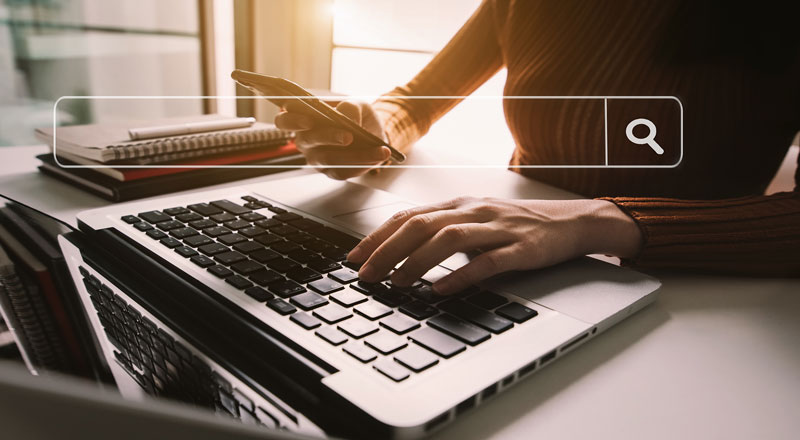 An image of someone using a laptop to search for information. They also have a phone. There is a search bar graphic towards the top of the image