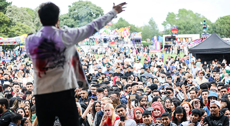 A photograph of a man singing from a stage. There is a large crowd in front of him and fairground rides are in the background.