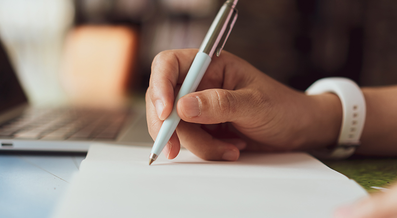 Person writing in a notebook (image shows hand, pen, notebook and laptop)