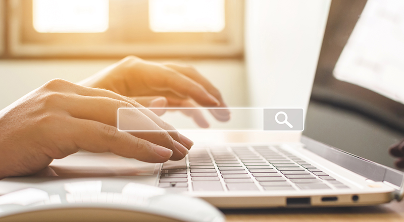 Person using a laptop (image shows laptop, mouse, hands in the background with a search bar in the foreground)