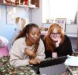 Two female students lie on a bed in their  room with a tablet.