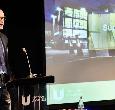 A man delivering a speech next to a PowerPoint presentation