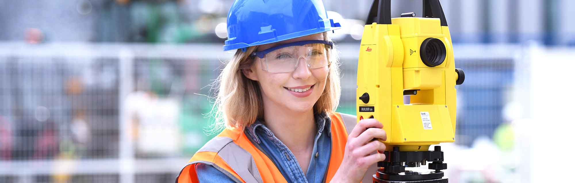 Female civil engineering student in PPE using theodolite on construction site