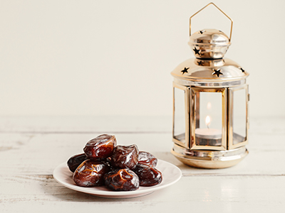 Pile of dried dates on white plate and gold coloured metal lantern with burning candle on wooden table.. Link to Shared Iftar – March 2026.