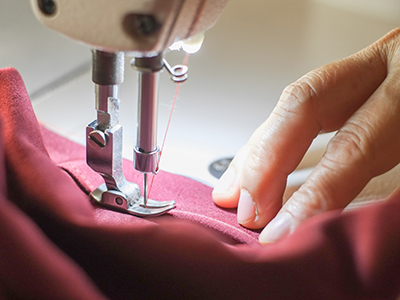 Someone using a sewing machine to hem a  garment - photo shows hand, fabric and sewing machine foot/ needle.. Link to Someone using a sewing machine to hem a  garment - photo shows hand, fabric and sewing machine foot/ needle..