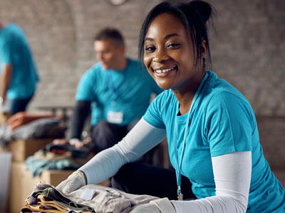 An image of a volunteer carrying clothes and smiling. Other volunteers can be seen in the background. . Link to Do Good Feel Good! Volunteering Opportunities - June 2026.