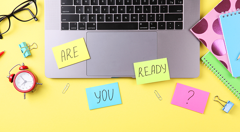 Open laptop, glasses, alarm clock, notebooks, paperclips and bulldog clip with post-it notes that have the words 'are you ready?' written on them.