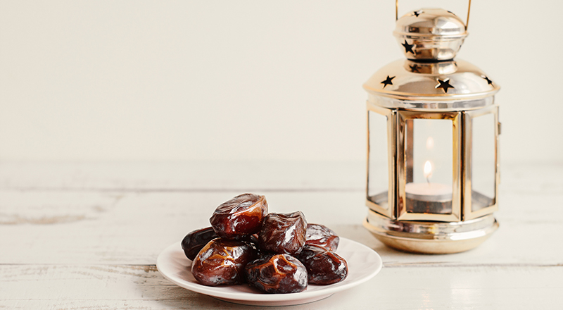 Pile of dried dates on white plate and gold coloured metal lantern with burning candle on wooden table.