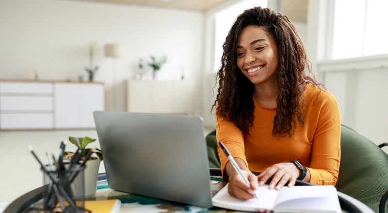 An image of a student writing in a notebook and working on a laptop.