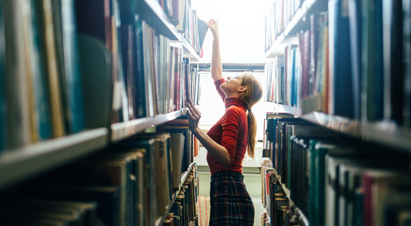 An image of a person reaching upwards to retrieve books from a book shelf
