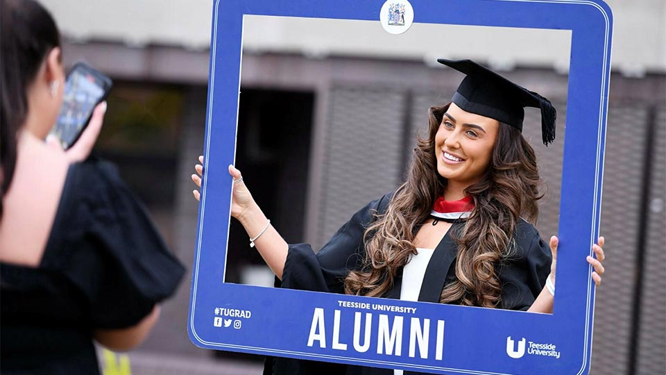 Graduate holding a seflie frame on campus