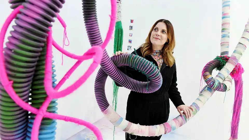Fine art student with their work at the Degree showcase. The work contains a series of coloured tubes in a random configuration.