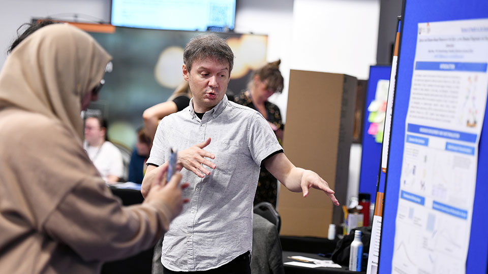 University students and staff engaging at a research showcase event. A university staff member explains a research poster to a student during an academic event, with other participants visible in the background.