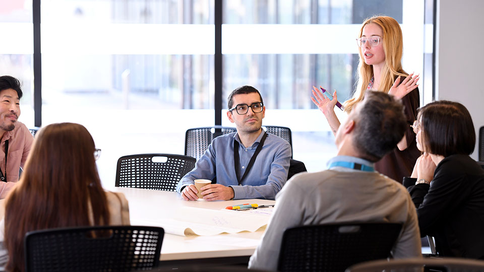 Staff sit in a table in a workshop with a women stand up talking