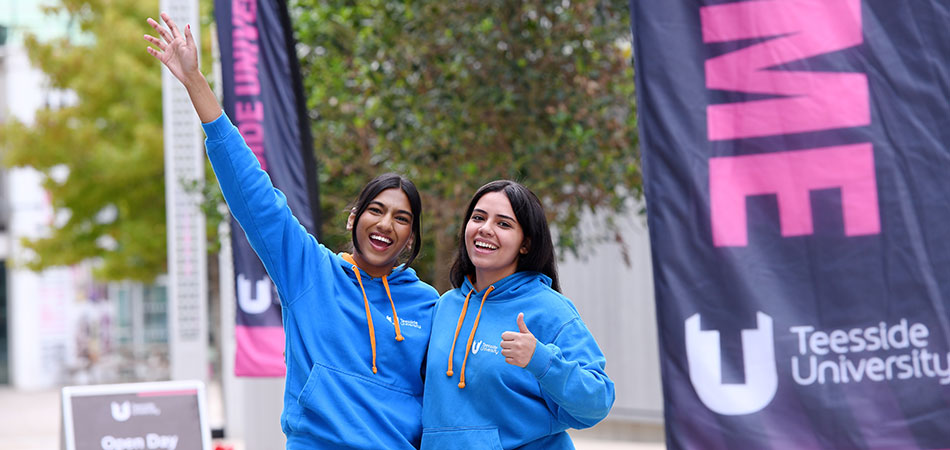 Two Teesside University student ambassadors smiling and waving in blue hoodies on campus near a Teesside University banne
