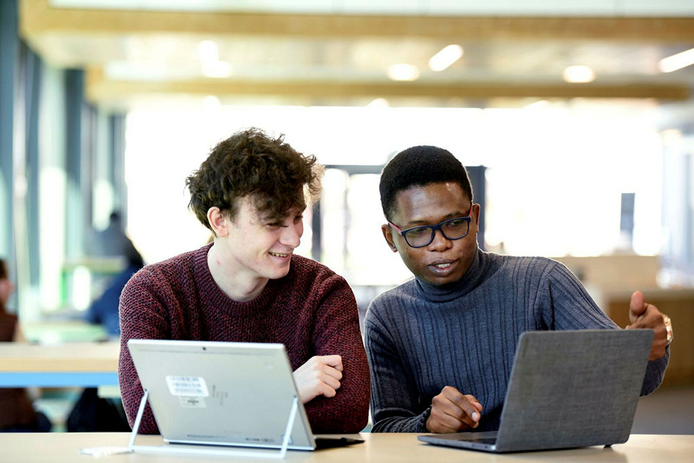 Two students seated side-by-side, each using a laptopâ€”one pointing at their screen while the other looks on.