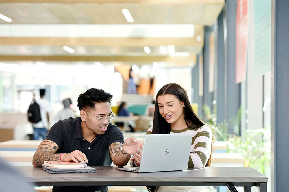 Two students sitting at a table, looking at a laptop togetherâ€”one student pointing at the screen while the other smiles.